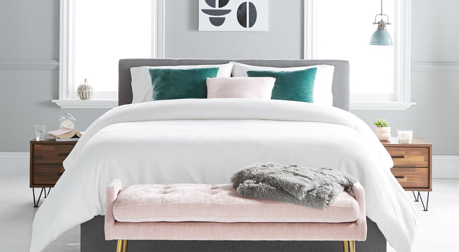 A bedroom styled with a gray upholstered mid-century style bed, two wooden mid-century style nightstands and a pink velvet upholstered bench and velvet decorative pillows. Links to where to buy luxury mid-century modern bedroom furniture and decor